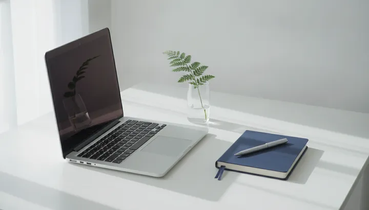 An open laptop, a sprig of fern in a glass and a notebook on a white desk